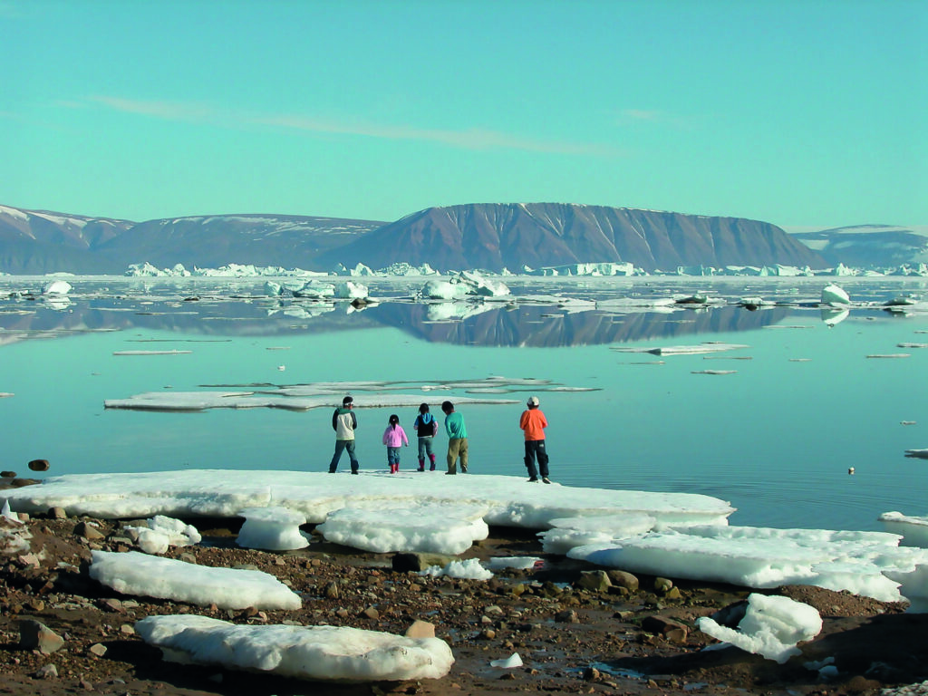 A group of five children playing at the beach, which still is covered with ice in places. They're facing the water, full of chunks of sea ice