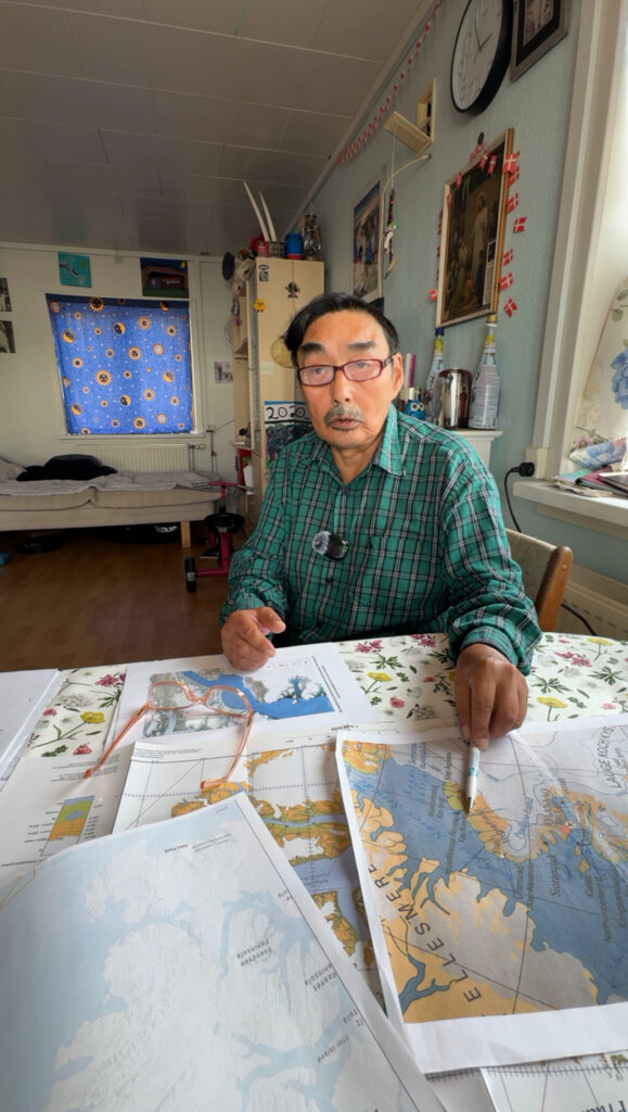 Man in green shirt sitting by a table with various maps in front of him.